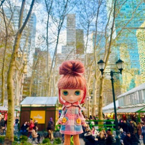 May include: A doll wearing a pink and blue plaid coat with yellow flower accents. The doll has red hair in a bun and blue eyes. The doll is standing in front of a city park with tall buildings in the background.