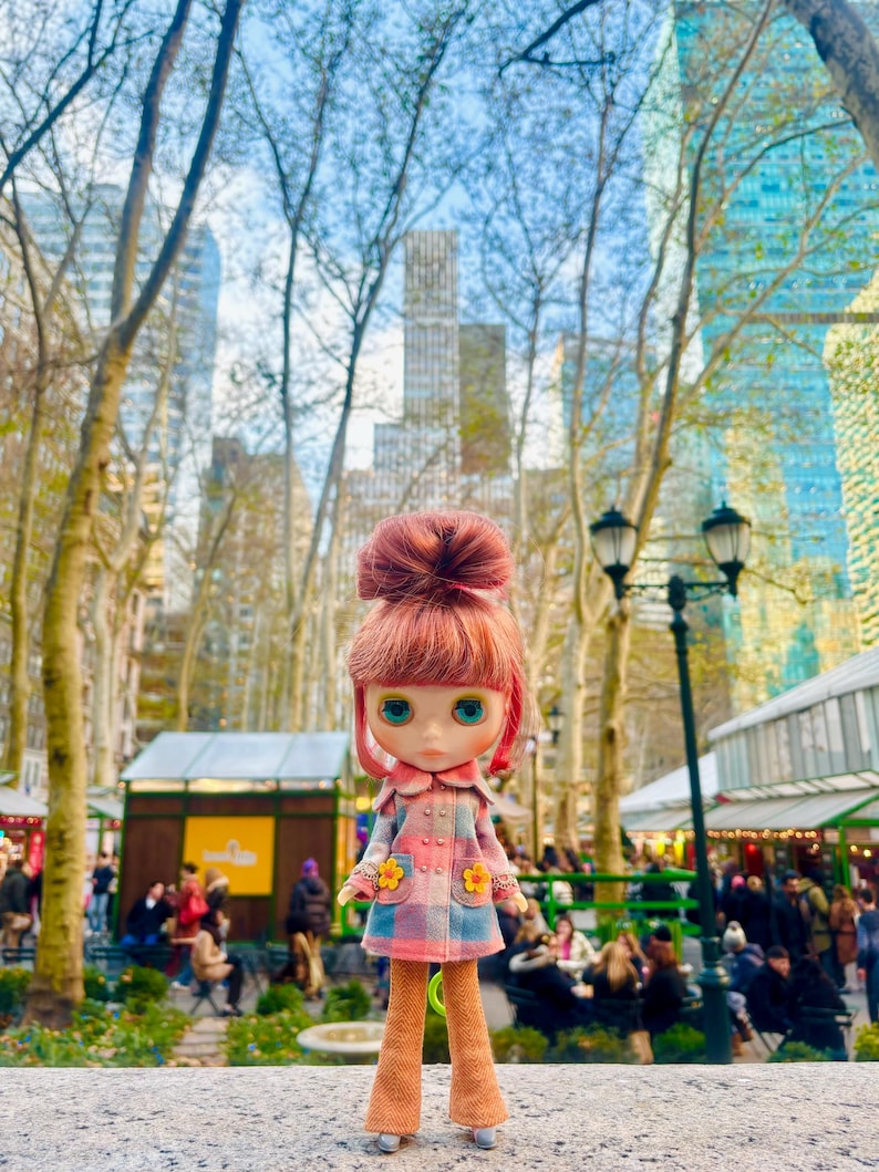 May include: A doll wearing a pink and blue plaid coat with yellow flower accents. The doll has red hair in a bun and blue eyes. The doll is standing in front of a city park with tall buildings in the background.