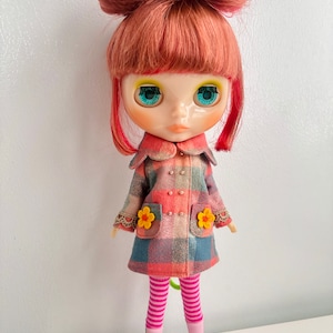 May include: A doll with red hair styled in a bun, wearing a plaid coat with flower accents, pink and white striped tights, and pink shoes.
