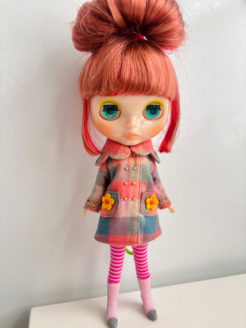 May include: A doll with red hair styled in a bun, wearing a plaid coat with flower accents, pink and white striped tights, and pink shoes.