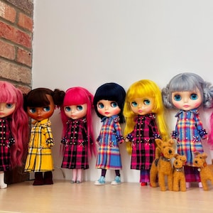 May include: A collection of eight dolls with diverse hair colors including red, pink, brown, black, yellow, and gray, all wearing long-sleeved coats in plaid patterns of yellow, pink, or blue. Three small brown dog figurines stand among them.