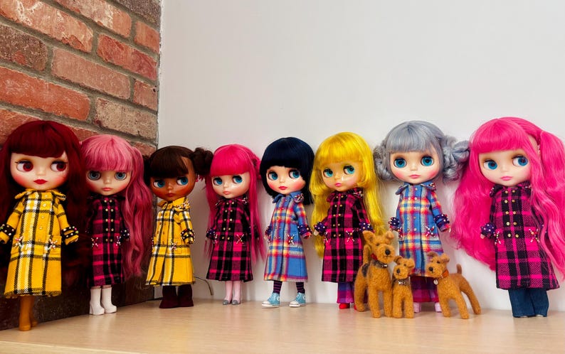 May include: A collection of eight dolls with diverse hair colors including red, pink, brown, black, yellow, and gray, all wearing long-sleeved coats in plaid patterns of yellow, pink, or blue. Three small brown dog figurines stand among them.