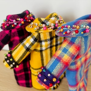 May include: Three miniature coats in plaid patterns of pink and black, yellow and black, and blue and red. Each coat features a wide collar and cuffs embellished with small, colorful beads in shades of pink, blue, yellow, and white, arranged in a decorative row.