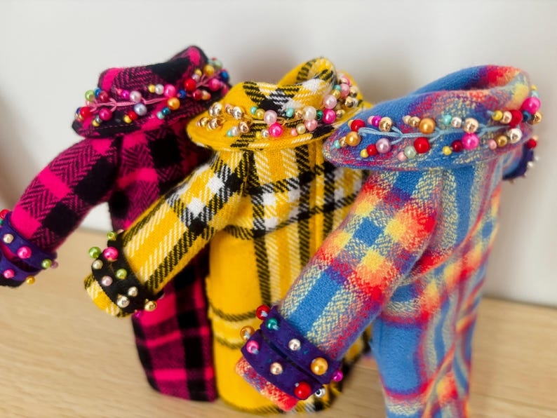May include: Three miniature coats in plaid patterns of pink and black, yellow and black, and blue and red. Each coat features a wide collar and cuffs embellished with small, colorful beads in shades of pink, blue, yellow, and white, arranged in a decorative row.