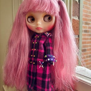 May include: A doll with long, straight pink hair and bangs, wearing a black and magenta plaid coat with gold buttons. The coat features decorative colorful beads on the cuffs and a small beaded cross detail on the front pocket area.