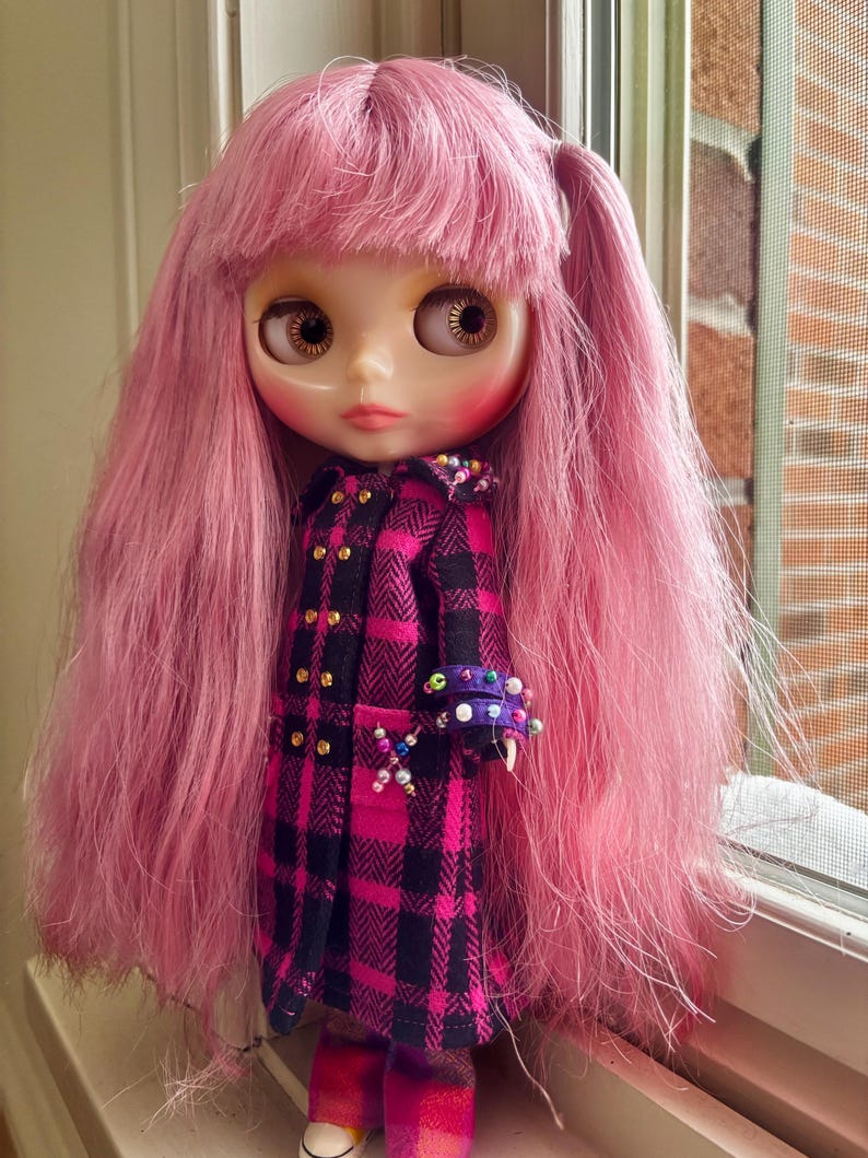 May include: A doll with long, straight pink hair and bangs, wearing a black and magenta plaid coat with gold buttons. The coat features decorative colorful beads on the cuffs and a small beaded cross detail on the front pocket area.