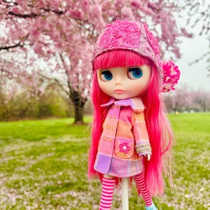 May include: A pink doll with long pink hair wearing a pink and purple plaid coat, a pink hat with lace detail, and blue and white striped socks. The doll is standing on a white stick in a field of pink flowers.
