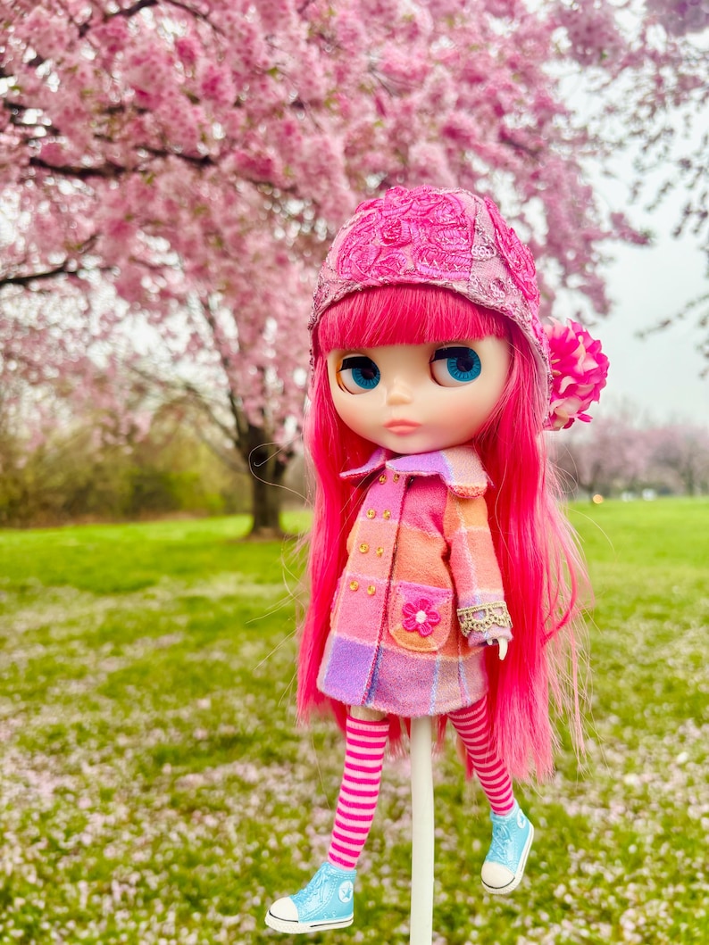 May include: A pink doll with long pink hair wearing a pink and purple plaid coat, a pink hat with lace detail, and blue and white striped socks. The doll is standing on a white stick in a field of pink flowers.