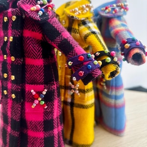 May include: Three miniature coats in pink, yellow, and blue plaid patterns. Each coat features gold-tone buttons, decorative beaded accents on the collars and cuffs, and a cross-shaped beaded design on the pockets. The coats are arranged in a row.