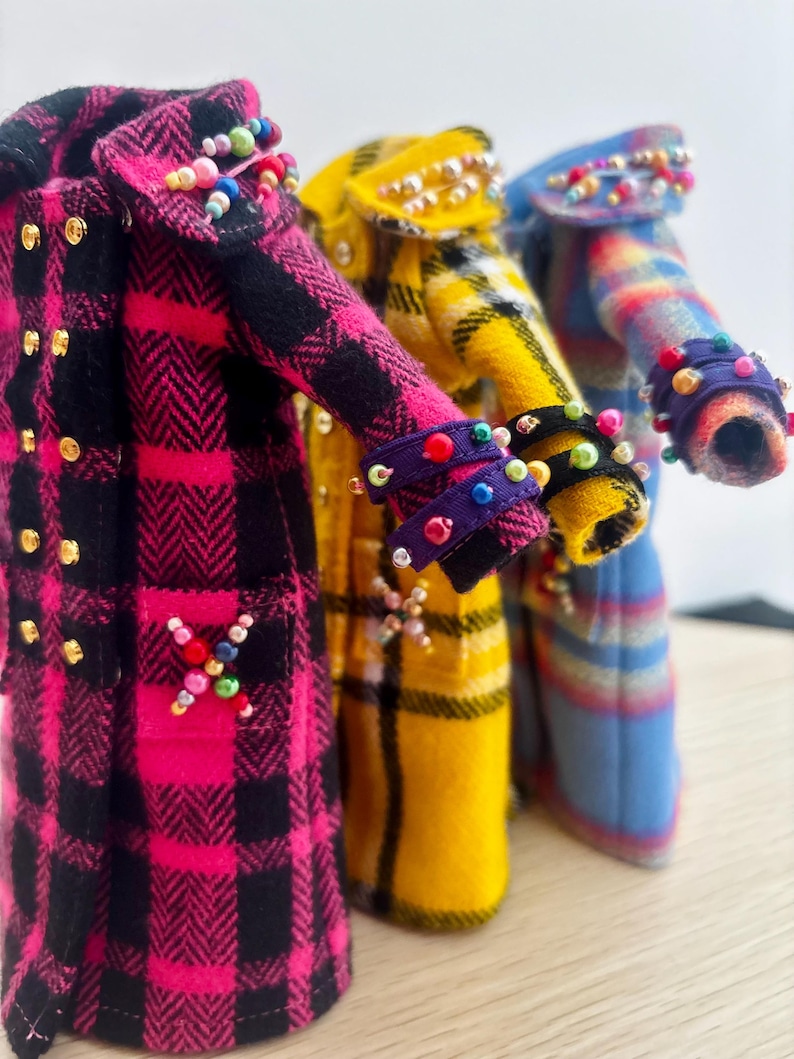 May include: Three miniature coats in pink, yellow, and blue plaid patterns. Each coat features gold-tone buttons, decorative beaded accents on the collars and cuffs, and a cross-shaped beaded design on the pockets. The coats are arranged in a row.