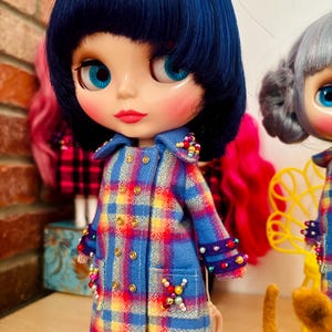 May include: A doll with short dark blue hair and bright blue eyes wearing a double-breasted blue, yellow, and red plaid coat. The coat features gold-tone buttons, purple cuffs, and decorative clusters of colorful beads on the collar and pockets.