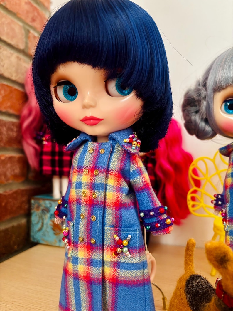 May include: A doll with short dark blue hair and bright blue eyes wearing a double-breasted blue, yellow, and red plaid coat. The coat features gold-tone buttons, purple cuffs, and decorative clusters of colorful beads on the collar and pockets.