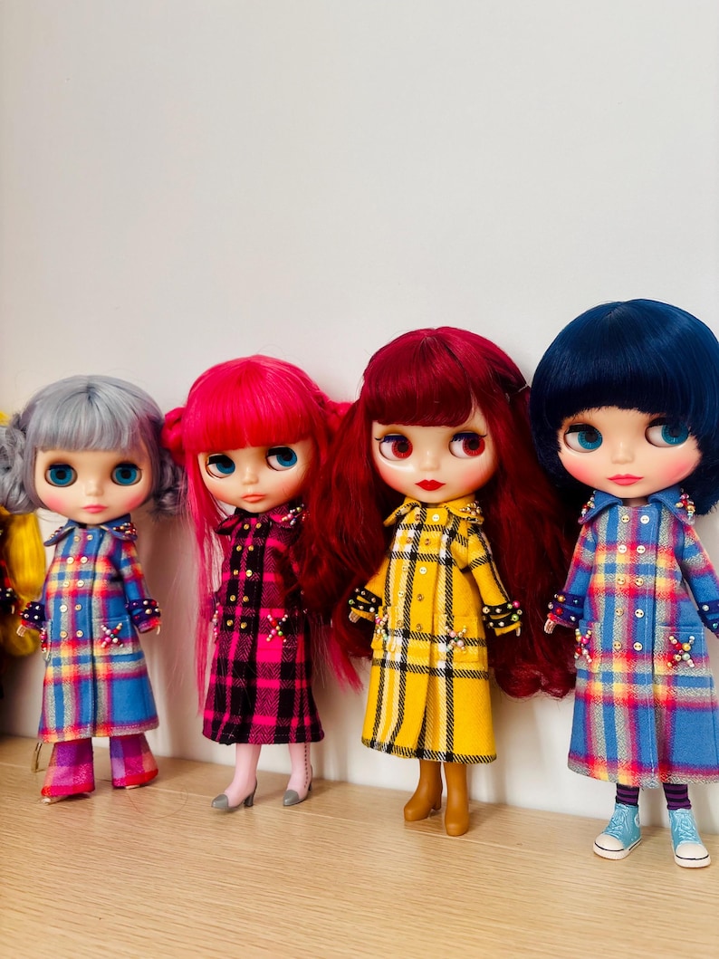 May include: Four dolls with large eyes and distinct hair colors, including silver, pink, red, and dark blue. Each doll wears a long-sleeved, button-down plaid coat featuring decorative bead accents on the pockets and cuffs, paired with matching footwear.