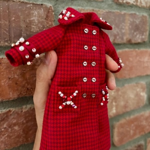 May include: A miniature red and dark red houndstooth coat featuring a double-breasted front with silver buttons, a wide collar, and long sleeves. The garment is embellished with clusters of white and red beads on the collar, cuffs, and pockets.