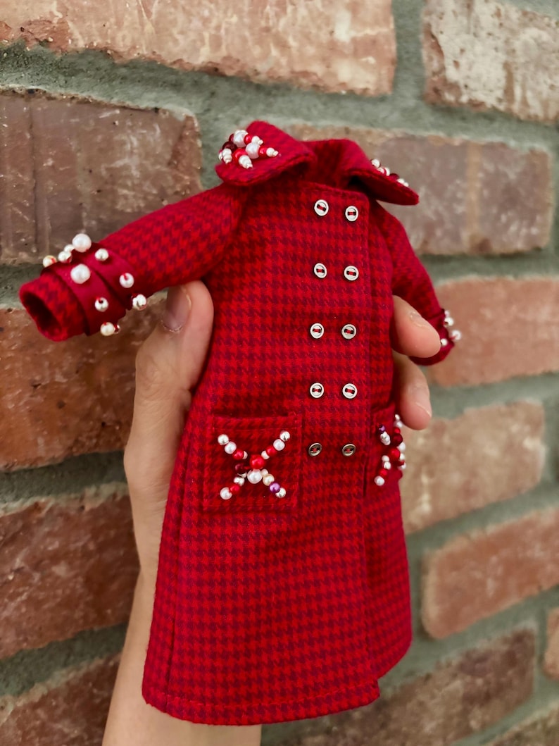 May include: A miniature red and dark red houndstooth coat featuring a double-breasted front with silver buttons, a wide collar, and long sleeves. The garment is embellished with clusters of white and red beads on the collar, cuffs, and pockets.