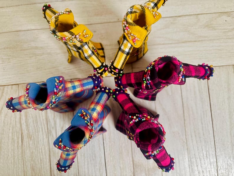 May include: Five miniature jackets arranged in a circle, featuring two yellow plaid, two pink and black plaid, and one blue and yellow plaid design. Each jacket is embellished with small, colorful beads along the collars and sleeve cuffs.