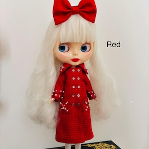 May include: A doll with long white hair and a large red bow, wearing a red plaid double-breasted coat embellished with small white beads on the collar, cuffs, and pockets. The doll has blue eyes, red lips, and white boots. The text Red is visible.