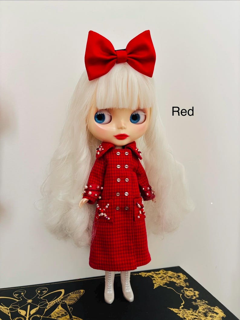 May include: A doll with long white hair and a large red bow, wearing a red plaid double-breasted coat embellished with small white beads on the collar, cuffs, and pockets. The doll has blue eyes, red lips, and white boots. The text Red is visible.