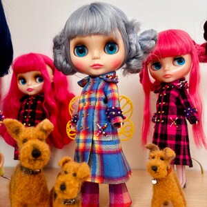 May include: Three dolls with large eyes and colorful hair, wearing plaid coats with beaded accents, stand alongside three small, brown, textured dog figurines. The central doll has silver hair, while the two flanking dolls have bright pink hair.