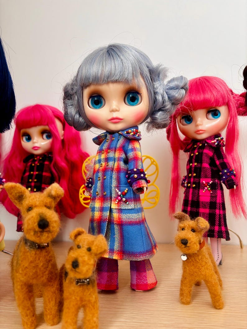 May include: Three dolls with large eyes and colorful hair, wearing plaid coats with beaded accents, stand alongside three small, brown, textured dog figurines. The central doll has silver hair, while the two flanking dolls have bright pink hair.