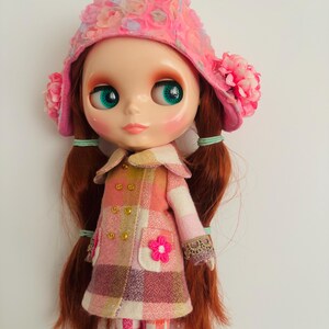 May include: A doll with long red hair wearing a pink and white plaid coat, a pink hat with flower embellishments, and pink and white plaid pants.