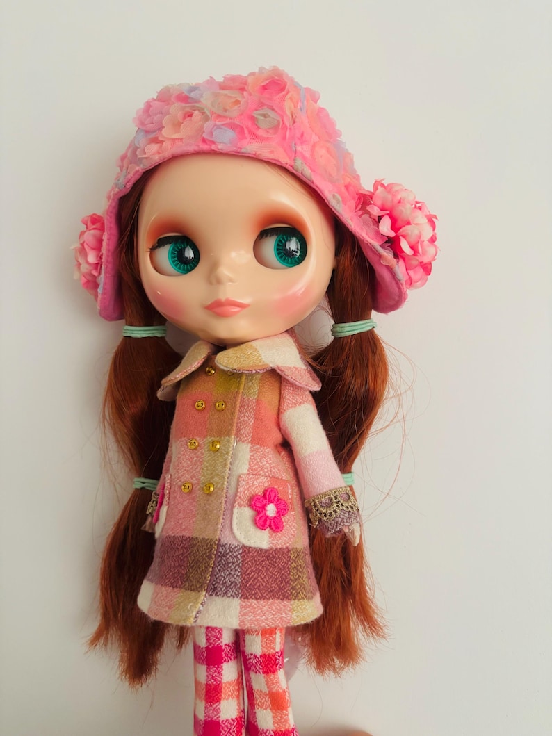 May include: A doll with long red hair wearing a pink and white plaid coat, a pink hat with flower embellishments, and pink and white plaid pants.