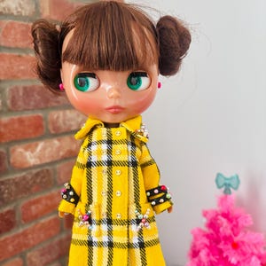 May include: A doll with brown hair styled in two buns and bangs, featuring green eyes and pink earrings. The doll wears a yellow, black, and white plaid double-breasted coat with small buttons, black cuffs adorned with colorful beads, and matching beaded accents on the pockets.