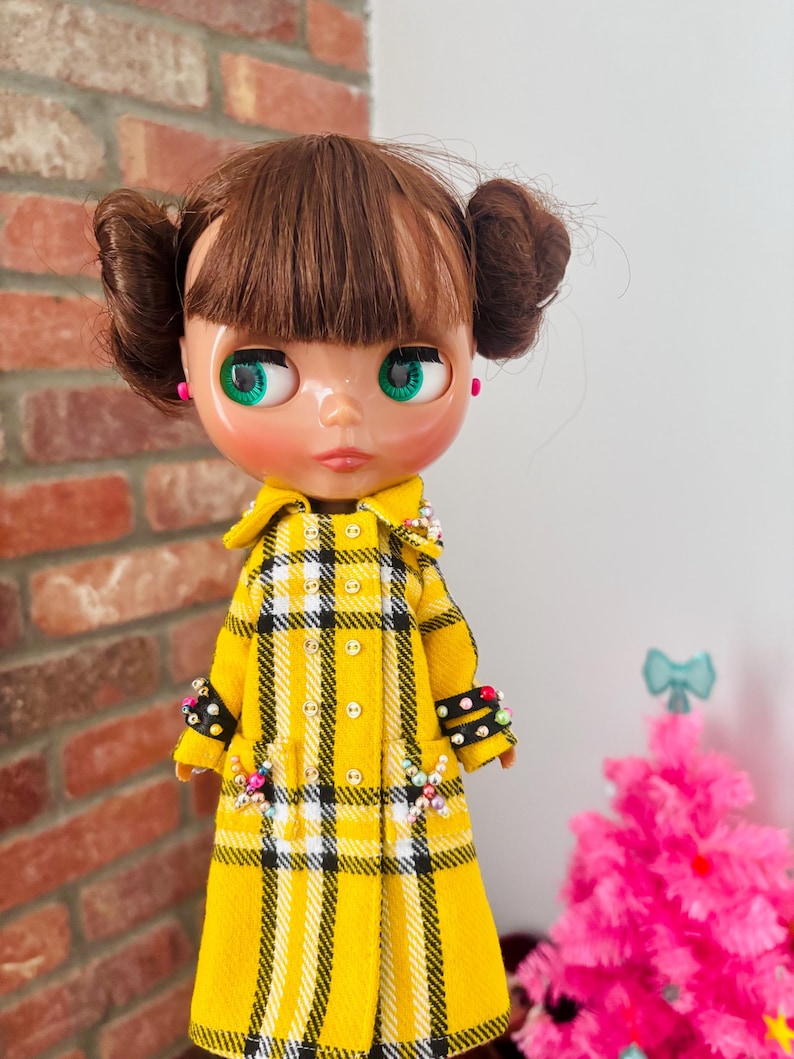 May include: A doll with brown hair styled in two buns and bangs, featuring green eyes and pink earrings. The doll wears a yellow, black, and white plaid double-breasted coat with small buttons, black cuffs adorned with colorful beads, and matching beaded accents on the pockets.