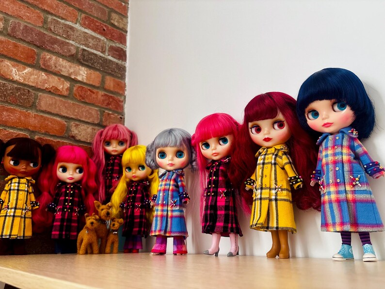 May include: A collection of eight dolls standing in a row, wearing various plaid coats in yellow, blue, and pink. The dolls feature diverse hair colors including pink, yellow, silver, red, and dark blue, and are positioned on a flat surface.