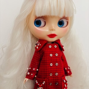 May include: A doll with long white hair and a red bow, featuring bright blue eyes and red lips. The doll wears a red and black houndstooth double-breasted coat adorned with white and red beads on the collar, cuffs, and pockets.