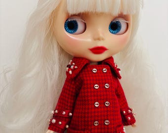 Può includere: Una bambola con lunghi capelli bianchi e un fiocco rosso, occhi blu brillante e labbra rosse. La bambola indossa un cappotto doppiopetto a motivo pied-de-poule rosso e nero, decorato con perline bianche e rosse su colletto, polsini e tasche.