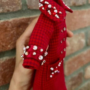 May include: A red and dark red houndstooth coat featuring a double-breasted front with small silver buttons. The collar and cuffs are embellished with white and red beads. A small pocket on the front is also decorated with a cluster of beads.