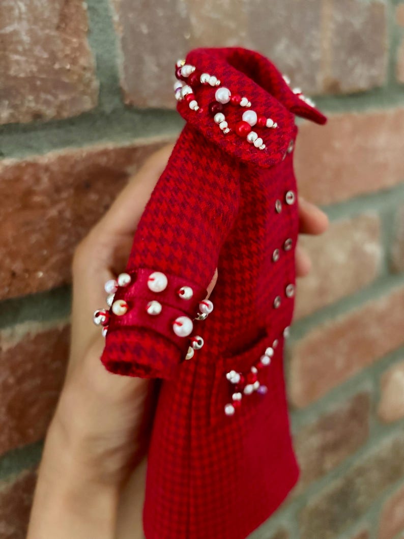 May include: A red and dark red houndstooth coat featuring a double-breasted front with small silver buttons. The collar and cuffs are embellished with white and red beads. A small pocket on the front is also decorated with a cluster of beads.