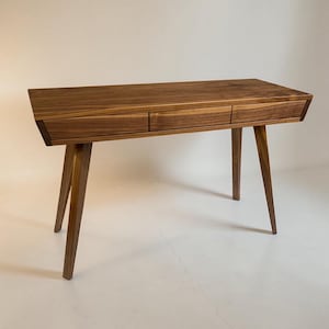 May include: A rectangular wooden desk featuring a rich brown grain pattern, three horizontal drawers aligned across the front, and four tapered legs that angle outward from the base to provide structural support for the flat tabletop surface.