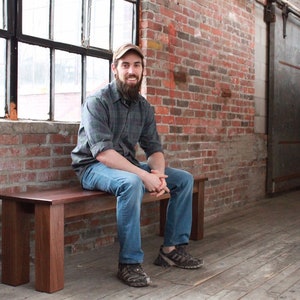 May include: A man with a beard sits on a wooden bench in a brick room. The bench is made of dark wood and has a simple design. The man is wearing a plaid shirt, blue jeans, and a baseball cap.
