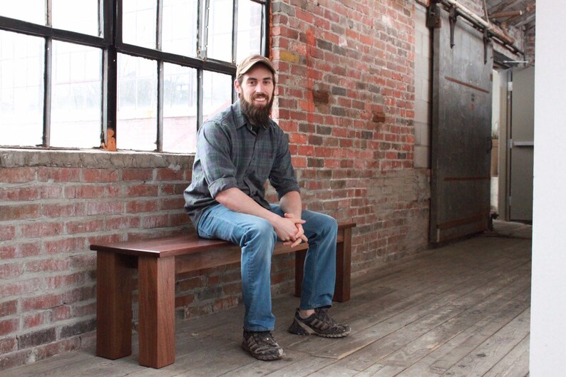 May include: A man with a beard sits on a wooden bench in a brick room. The bench is made of dark wood and has a simple design. The man is wearing a plaid shirt, blue jeans, and a baseball cap.