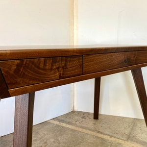 May include: A dark brown wooden desk featuring a smooth rectangular top and three integrated drawers across the front. The desk is supported by four angled, tapered legs that extend outward from the base, showcasing a natural wood grain finish.