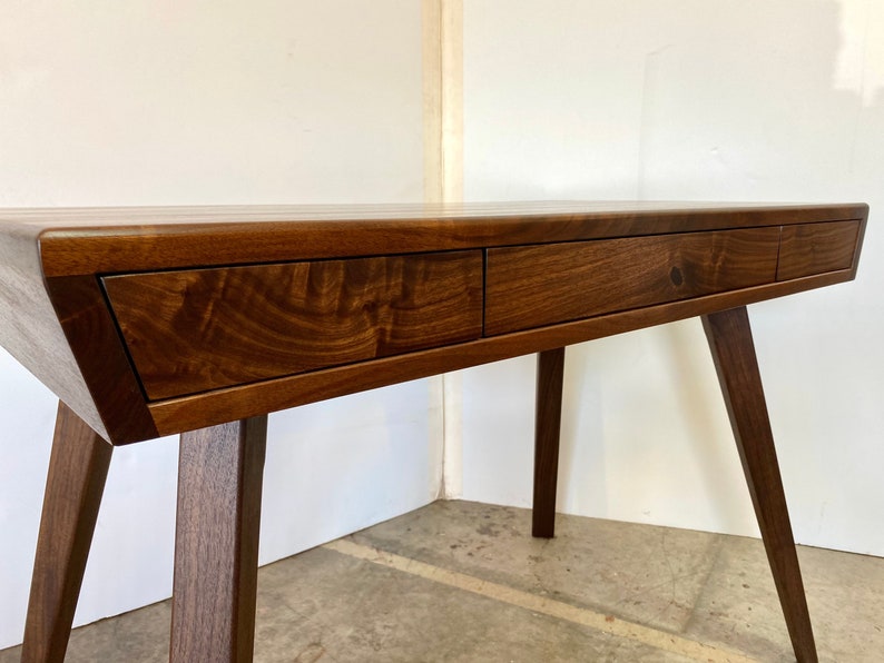 May include: A dark brown wooden desk featuring a smooth rectangular top and three integrated drawers across the front. The desk is supported by four angled, tapered legs that extend outward from the base, showcasing a natural wood grain finish.