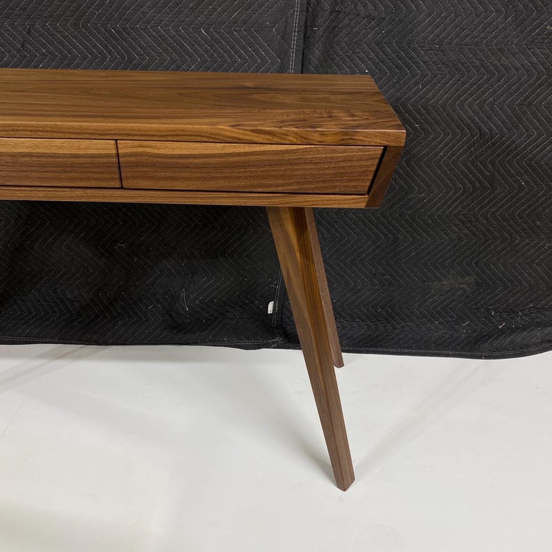May include: A brown wooden console table featuring a smooth rectangular top and two integrated drawers with a horizontal wood grain pattern. The table is supported by angled, tapered legs that extend outward from the base of the frame.