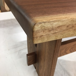 May include: A wooden bench with a live edge top and a natural wood finish. The bench has a simple design with a single support beam on each side.