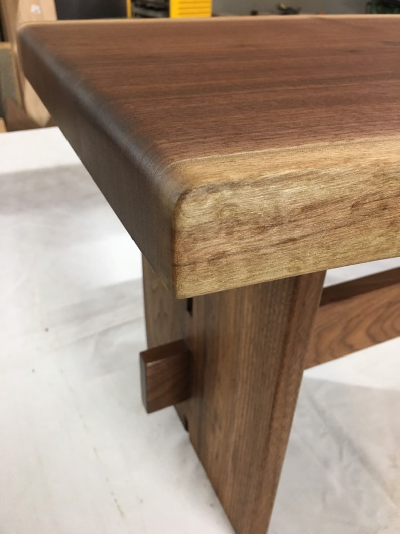 May include: A wooden bench with a live edge top and a natural wood finish. The bench has a simple design with a single support beam on each side.