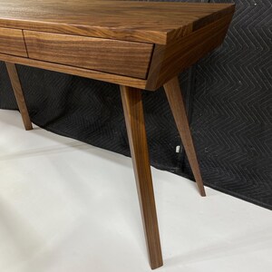 May include: A dark brown wooden console table featuring a smooth rectangular top, two integrated drawers with horizontal grain patterns, and four tapered, angled legs that extend outward from the base to provide a stable foundation.