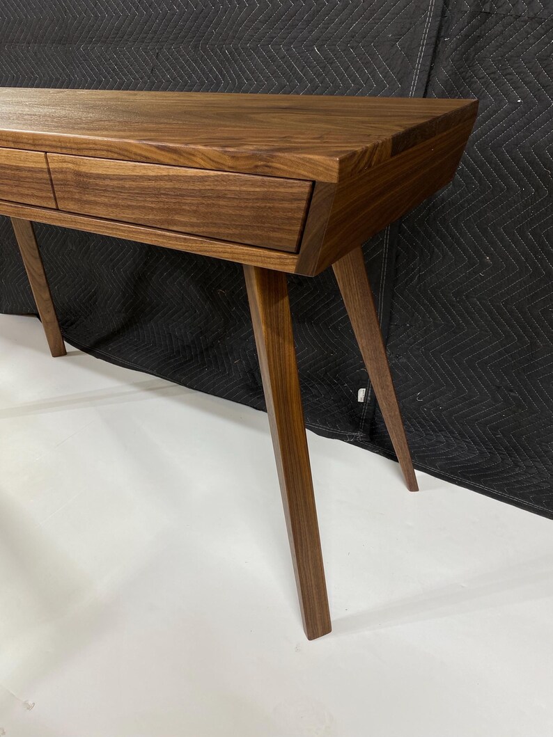 May include: A dark brown wooden console table featuring a smooth rectangular top, two integrated drawers with horizontal grain patterns, and four tapered, angled legs that extend outward from the base to provide a stable foundation.
