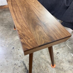 May include: A rectangular wooden desk featuring a rich brown walnut grain pattern on the flat top surface. The desk is supported by four tapered wooden legs, each finished with a small, bright orange circular protective cap at the base.