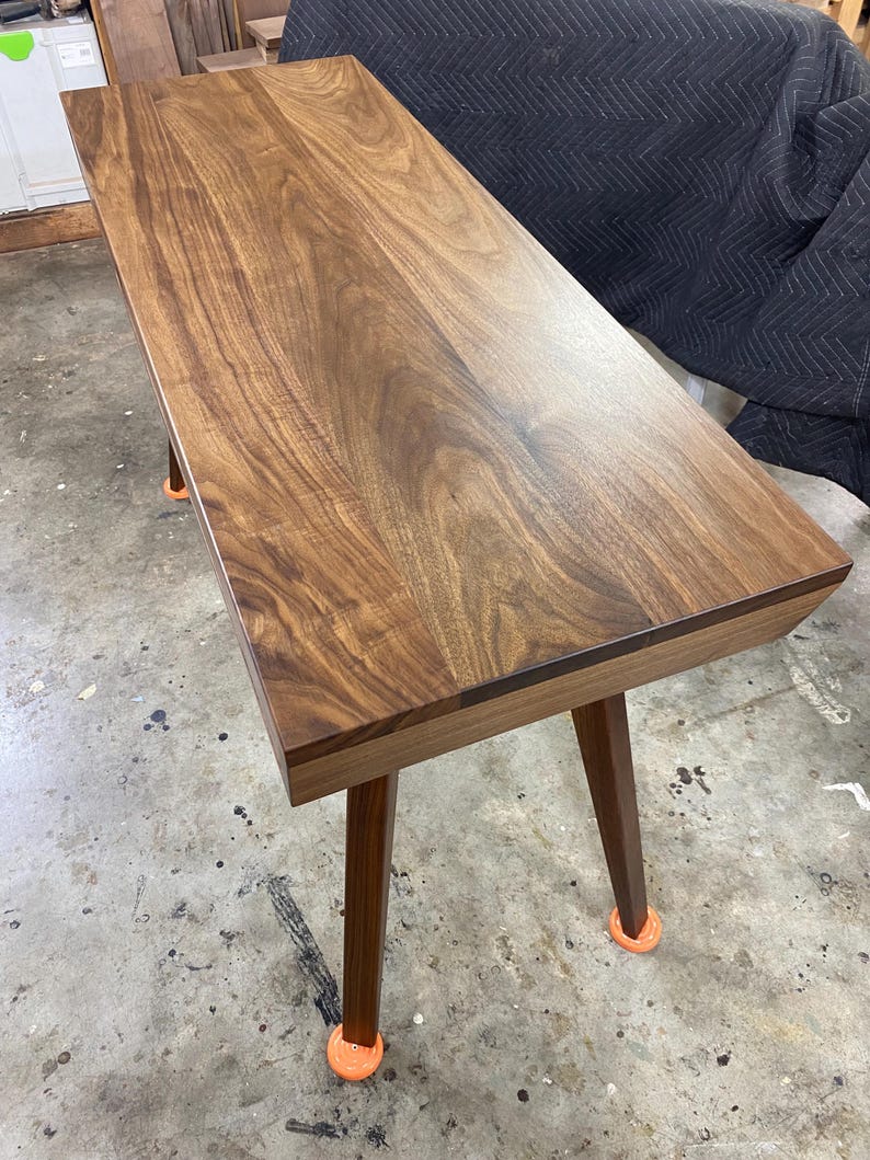 May include: A rectangular wooden desk featuring a rich brown walnut grain pattern on the flat top surface. The desk is supported by four tapered wooden legs, each finished with a small, bright orange circular protective cap at the base.
