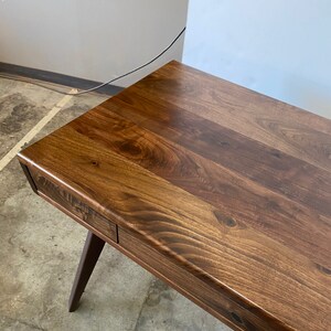May include: A dark brown wooden desk featuring a smooth, polished surface with visible natural wood grain patterns and knots. The desk includes a single integrated drawer along the front edge and is supported by a tapered, angled wooden leg.