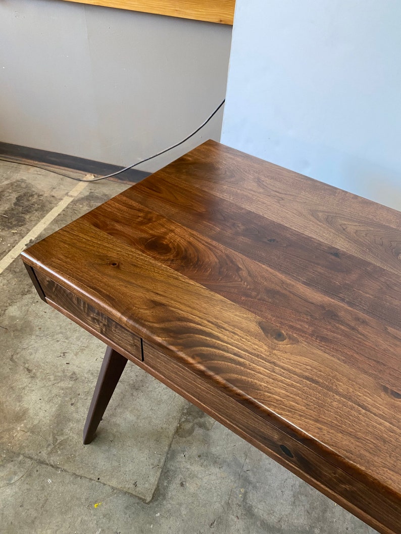 May include: A dark brown wooden desk featuring a smooth, polished surface with visible natural wood grain patterns and knots. The desk includes a single integrated drawer along the front edge and is supported by a tapered, angled wooden leg.