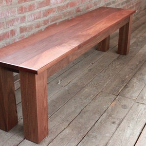 May include: A long, wooden bench with a natural reddish-brown finish. The bench has a simple, rectangular design with four legs.