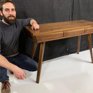May include: A rectangular wooden desk crafted from dark brown walnut wood. The desk features a smooth flat top surface and three horizontal drawers positioned side by side along the front. It is supported by four tapered legs angled outward.