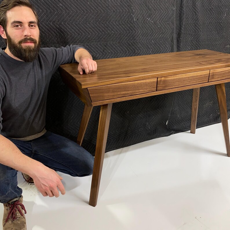 May include: A rectangular wooden desk crafted from dark brown walnut wood. The desk features a smooth flat top surface and three horizontal drawers positioned side by side along the front. It is supported by four tapered legs angled outward.